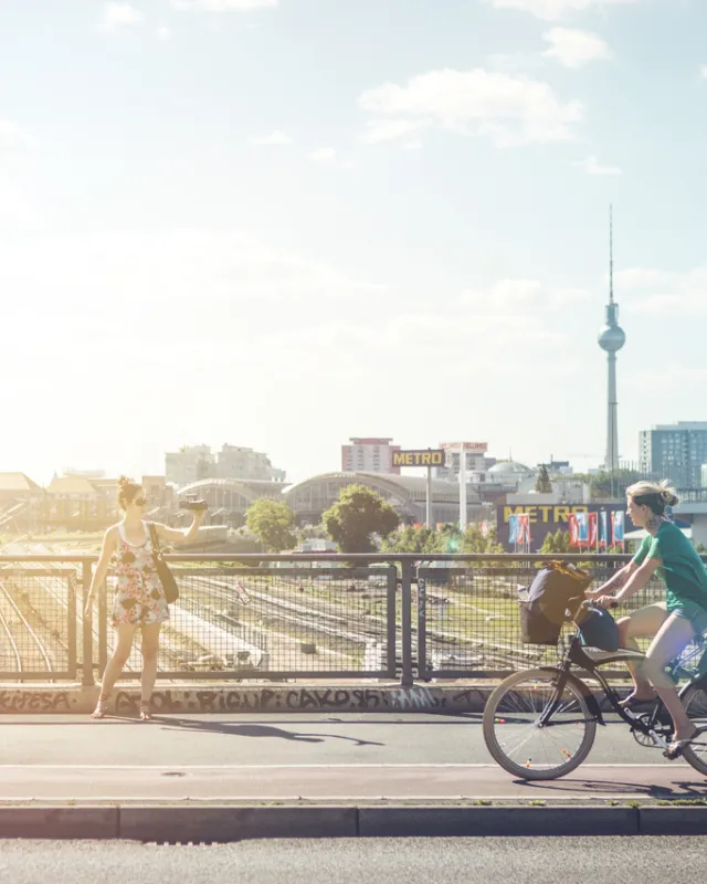 Frau auf Warschauer Brücke mit Kamera in Hand und Rücken zu TV Turm, andere Frau mit Fahrrad Richtung Oberbaumbrücke und Kind in Kindersitz, sonniges Licht